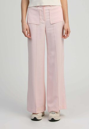 Femme portant un pantalon large rose pâle avec de grandes poches avant et des baskets beiges, debout devant un fond neutre.