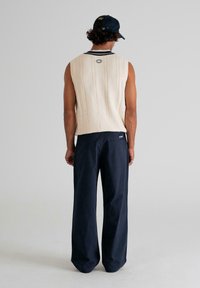 Man wearing a cream sleeveless cable-knit sweater, navy wide-leg pants, and a black cap, standing facing away against a plain background.