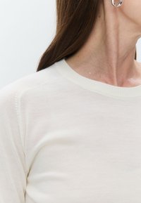 Pull en mailles crème clair avec un col rond, des bords côtelés et des détails de couture subtils. Texture de tissu douce et forme ajustée.