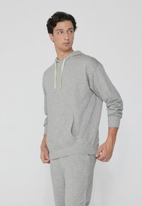 Grauer Hoodie aus weichem Stoff, mit Kängurutasche, verstellbarem Kapuzenzug und gerippten Bündchen. Trägt eine passende graue Hose.