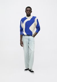 Jersey de punto en azul y beige con un patrón ondulado, combinado con pantalones vaqueros rectos azul claro y zapatillas negras con suelas blancas.