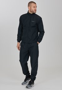 Svart vindjacka med dragkedja framtill och elastiska muddAR, kombinerat med matchande byxor. Har en logga på bröstet och en elegant design.