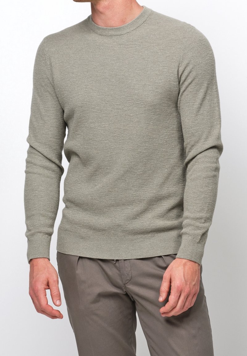 Homme portant un pull texturé gris clair à manches longues et un pantalon gris, visible de la poitrine jusqu'à la mi-cuisse sur un fond uni.