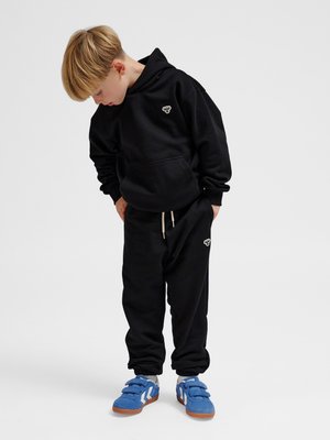 Młody chłopiec w czarnej bluzie z kapturem i spodniach dresowych patrzy w dół, nosi niebieskie trampki z białymi paskami, stoi na gładkim jasnym tle.