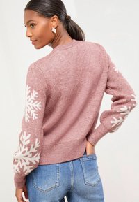 Rosa Pullover mit weißen Schneeflockenmuster an den Ärmeln, geripptem Saum und Bündchen. Getragen mit blauen Jeans. Hoher Rundhalsausschnitt und weiche Textur.
