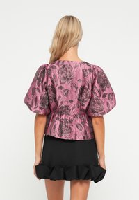 Rozā ziedu raksta peplum tops ar pūkainām piedurknēm, teksturēts audums un melna kroku svārki. Redzamie šuvumi un smalks spīdums materiālā.