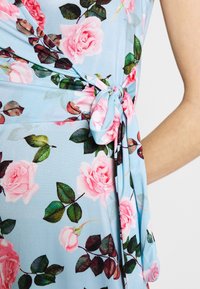 Robe bleu clair avec des motifs de roses roses et de feuilles vertes. Le tissu est lisse, nouée à la taille avec un détail noué. Design à manches courtes.