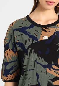 Czarny t-shirt z krótkim rękawem, ozdobiony zielonym i brązowym wzorem liści. Okrągły dekolt z małym detalem logo na piersi.