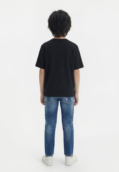 Παιδί με μαύρα μαλλιά που στέκεται με την πλάτη γυρισμένη, φοράει μαύρο μπλουζάκι, μπλε τζιν και λευκά αθλητικά παπούτσια, μπροστά σε απλό φόντο.