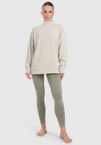 Femme debout pieds nus portant un pull à col roulé beige clair et des leggings verts délavés sur un fond uni.