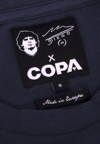 Zwarte kledinglabel met een portret, handtekening, "COPA," en "Made in Europe." Label is genaaid in een donkere stof. Maatlabel "S."