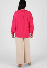 Blusa fucsia a maniche lunghe con collo arricciato, abbinata a pantaloni beige a gamba larga. Tessuto morbido con una leggera lucentezza.