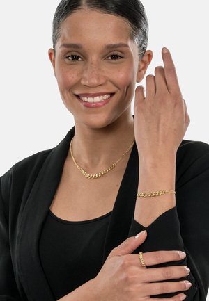 Goldene Halskette mit verknüpftem Design, passendes Armband und Ring, getragen über einem schwarzen Oberteil, welches die kettenartige Textur und den Glanz hervorhebt.