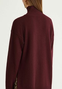 Maglione dolcevita color bordeaux in maglia strutturata, con collo e polsini a costine, vestibilità ampia e spacchi laterali per facilitare i movimenti.