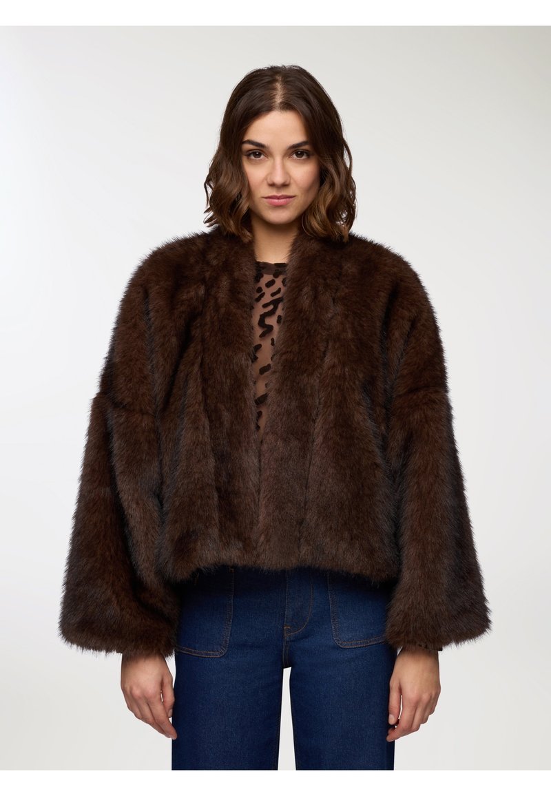 Giacca in pelliccia artificiale marrone con un design corto, maniche ampie e una texture morbida. Indossata sopra una maglietta trasparente con stampa leopardata e abbinata a jeans.