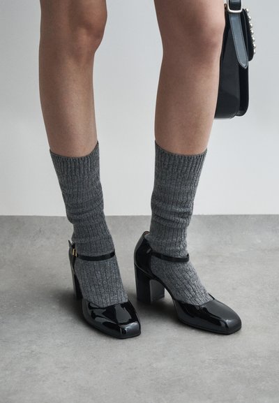 Sapatos de salto alto em pele estofada preta, com biqueira quadrada, usados com meias de lã acanaladas cinzas texturizadas. Apresentam detalhe de tira no tornozelo e salto bloco.