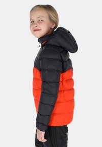 Jong kind met licht haar, gekleed in een zwart-oranje gewatteerde jas met capuchon, staand in profiel tegen een effen achtergrond.