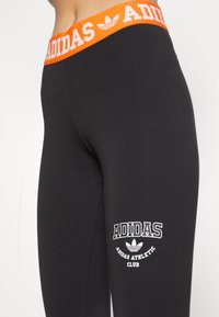Čierne legíny s kontrastným oranžovým elastickým pásom s bielym značkovým nápisom "ADIDAS". Obsahuje biele logo a textový detail na nohe.
