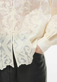 Broderet bluse i lys creme med intrikate blomster mønstre, en knaplukning og blødt plisserede ærmer; parret med sorte bukser.