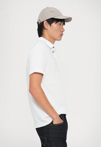 Hvid polo-shirt med krave og korte ærmer, parret med mørke jeans. En lysegrå kasket fuldender outfittet.