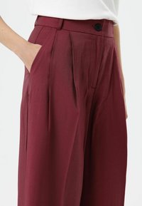 Bordeauxrode broek met plooien, gemaakt van lichtgewicht materiaal. Voorzien van zijzakken en een knoopsluiting in de taille. Gladde textuur.