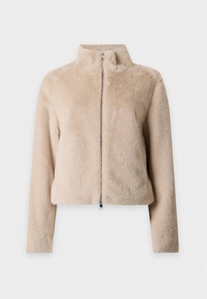Un mannequin se tient devant un fond uni, vêtu d’une veste en fausse fourrure rose pâle, d’un haut gris, d’un jean foncé et de bottines noires.
