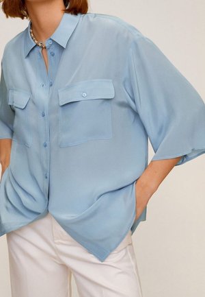 Femme portant une chemise ample bleu clair boutonnée avec deux poches poitrine et un pantalon blanc, les mains dans les poches, devant un fond uni.