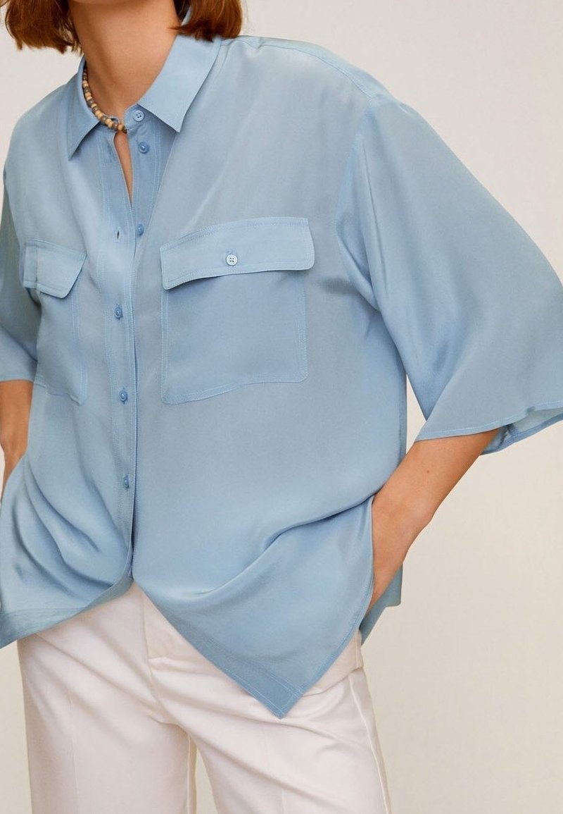 Femme portant une chemise ample bleu clair boutonnée avec deux poches poitrine et un pantalon blanc, les mains dans les poches, devant un fond uni.