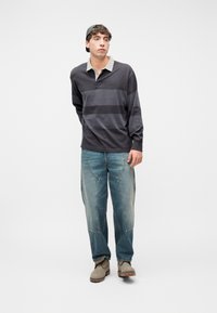 Jeune homme portant un maillot de rugby gris foncé à rayures, un jean en denim clair avec des patchs, une casquette grise portée à l'envers, et des bottes beiges sur un fond blanc.