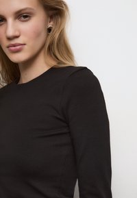 Črna majica z dolgimi rokavi in okroglim izrezom, narejena iz gladkega materiala. Ima diskretne šive in prilegajoč kroj.