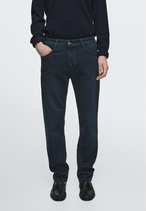 Persoon draagt donkerblauwe rechte jeans, zwarte loafers en een zwart geribd langarmshirt, met één hand in de zak.