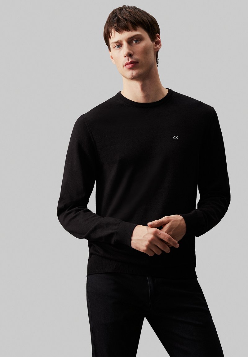 Svart langermet genser laget av mykt stoff, med rund hals og en liten hvit logo på brystet. Kombinert med mørke jeans.