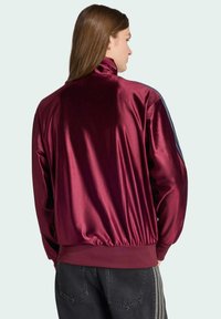 Vinrød satinjakke med høj krave, elastisk kant og kontrasterende blå striber på ærmerne, stylet med løse grå jeans.