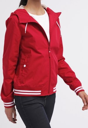 Persoon die een rood zip-up capuchonvest draagt met witte trekkers en gestreepte manchetten over een wit overhemd en donkere broek.