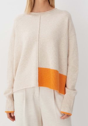 Mujer con un suéter beige extragrande con dobladillo y puños anchos acanalados en naranja, combinado con pantalones beige holgados.