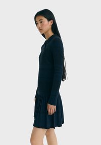 Nuori nainen, jolla on pitkät mustat hiukset, pukeutuneena tummansiniseen pitkään hihalliseen mekkoon, jossa on laskostettu helma, seisoo sivuttain vaaleaa taustaa vasten.