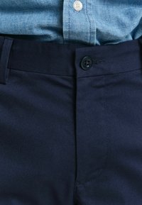 Marineblå bukser laget av glatt stoff, med knappelukking og flat frontdesign. Livet er pent avsluttet med søm.