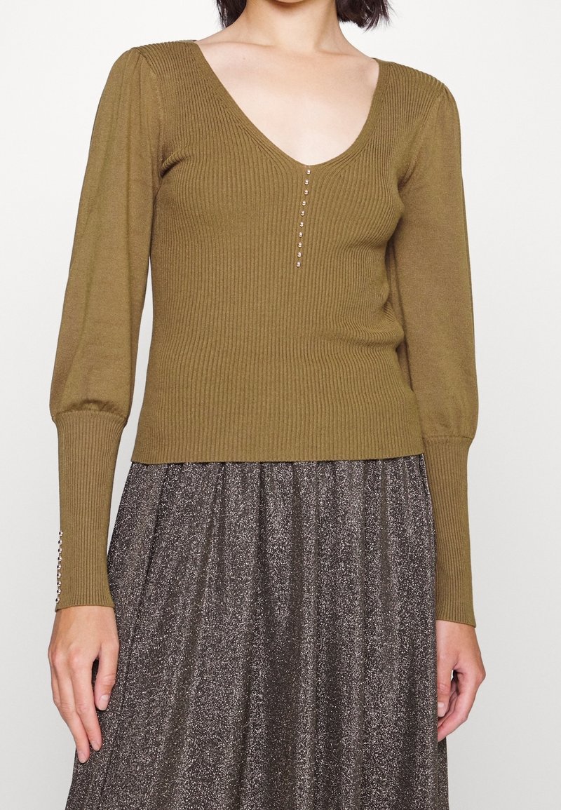 Olijfgroene geribbelde top met lange mouwen, V-hals en decoratieve knopen aan de voorkant; gecombineerd met een glanzende, donkergrijze rok.