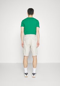 Zelené polo tričko, béžové cargo šortky, bílé ponožky a tenisky. Viditelné kapsy na šortkách. Neutrální pozadí.