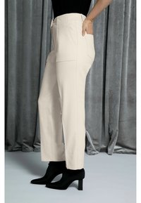 Béžové kalhoty na míru se rovnými nohavicemi, vysokým pasem a bočními kapsami, doplněné černými koženými kotníkovými botami na podpatku na šedém pozadí.