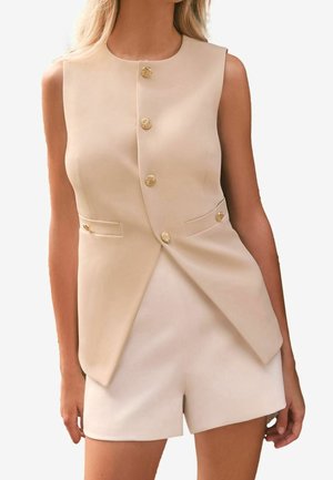 Blazer beige sans manches avec boutons dorés, associé à un short beige taille haute assorti porté par une femme debout.