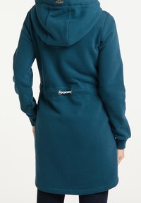 Cappotto lungo verde acqua con cappuccio e vita aderente, con un accento in catena argentata. Tessuto morbido con polsini e orlo a coste.