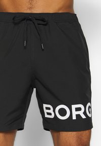 Juodos maudymosi šortai su elastingu juosmeniu ir raišteliu, su dideliu baltu tekstu "BORG" kairiajame šone perforuotu rašalu.