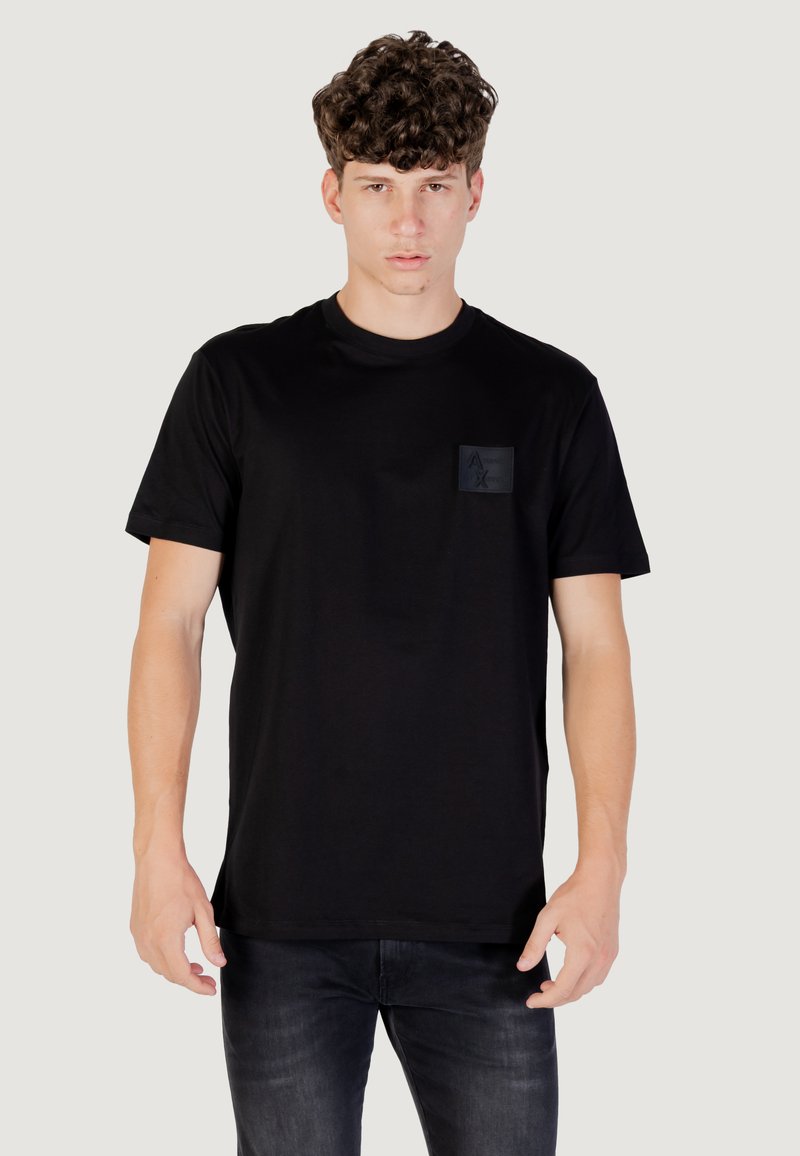 Camiseta negra de manga corta en tejido suave con un corte ajustado, que presenta un pequeño parche negro cuadrado en el pecho izquierdo con un detalle de logo.