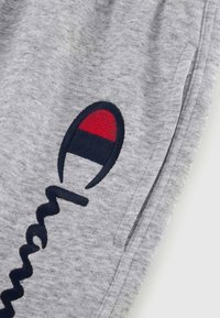 Graue Baumwoll-Sweatpants mit einem marineblauen gestickten Logo und roten sowie weißen Akzenten. Enthält eine Seitentasche und hat eine weiche Textur.