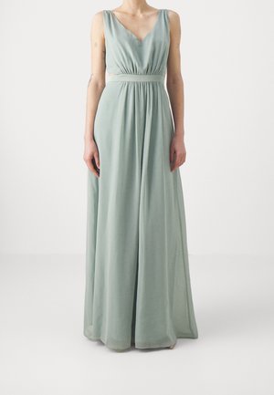 Femme portant une robe longue vert pâle sans manches avec un col en V et une taille froncée, debout devant un fond blanc uni.
