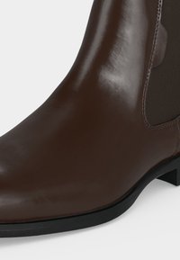 Gros plan d'une botte en cuir marron foncé brillant avec un panneau latéral élastique et un petit talon noir sur un fond clair uni.