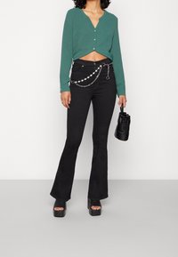 Cardigan corto verde con bottoni, jeans neri a campana con un accessorio a catena e sandali neri con platform. Piccola borsa nera in mano.