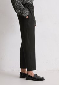 Persoon in zwarte cropped broek, zwarte gepatineerde loafers en een gestructureerde grijze trui met handen in de zakken tegen een witte muur.