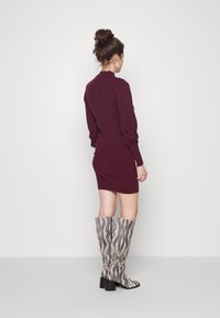 Abito lungo bordeaux con maniche lunghe, collo alto e silhouette aderente, abbinato a stivali al ginocchio con un motivo ondulato bianco e nero.